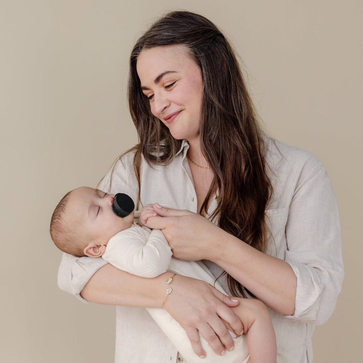 Woman holding a sleeping baby against a beige background, baby sucking on soothe beginnings pacifier